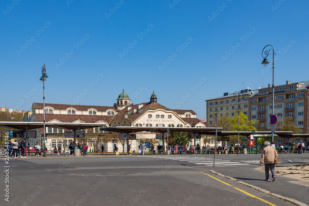 Outdoor sunny street view at bus station with people wait for buses, and background of Budova Městské tržnice, Municipal Market Building, against blue sky in Karlovy Vary, Czech Republic.