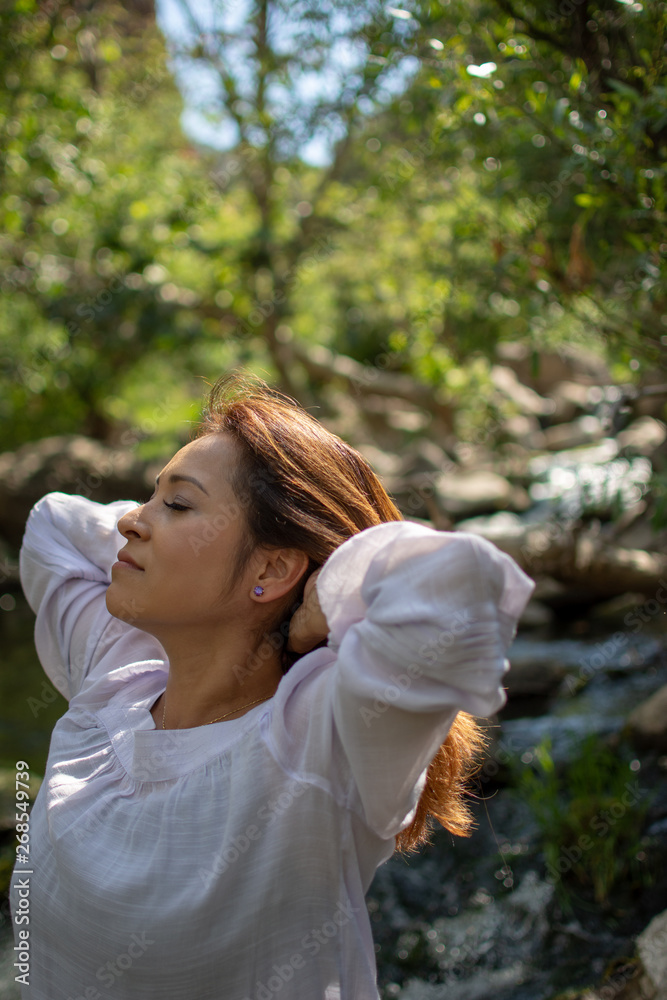 Naklejka premium Latina woman throwing her hair back with her hair in the sun in front of woods and a stream in the shade
