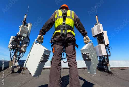 Professional industrial climber in helmet and uniform holds telecomunication equipment in his hand and antennas of GSM DCS UMTS LTE bands, outdoor radio units, optic fibers, power cables are installed photo
