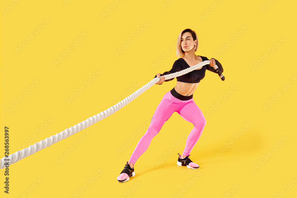 Strong woman pulling rope. Photo of sporty attractive woman in pink and black sportswear isolated on yellow background. Strength and motivation.