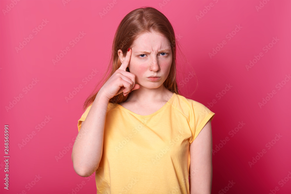 Image of focused young Caucasian girl touching temples with index fingers and frowning, has pensive facial expression, looks thoufhtfully directly at camera, wearing casually. Time to think concept. Image of focused young Caucasian girl touching temples with index fingers and frowning, has pensive facial expression, looks thoufhtfully directly at camera, wearing casually. Time to think concept.