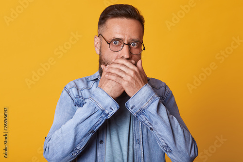 Shocked scared middle aged surprised face covering mouth with both hands, horrified stunned looking at camera isolated on yellow studio background, startled guy wearing denim kjacket and gray jacket. photo