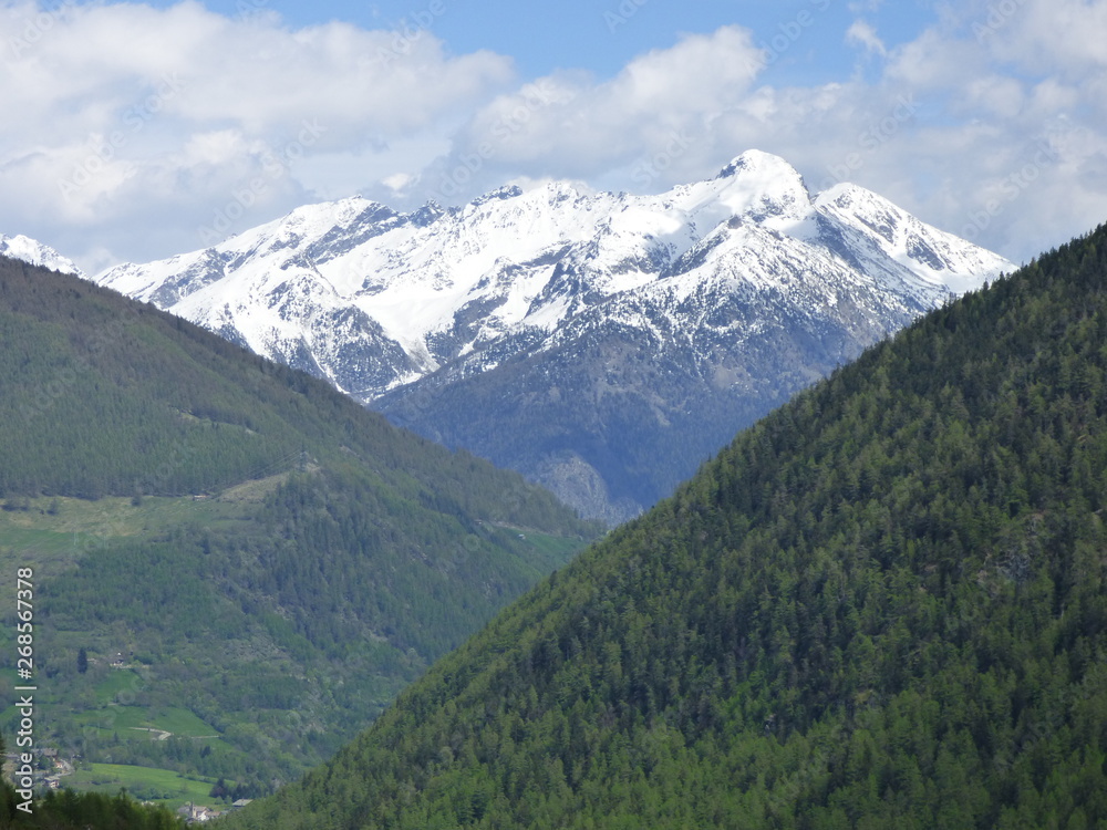 A snow capped peak of the Swiss Alps rising above lesser peaks and a mountain valley with clouds and a blue sky background