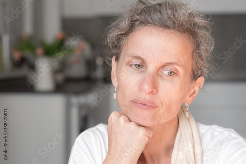 Closeup head and shoulders view of attractive middle aged woman resting head on hand and looking pensive with kitchen in background (selective focus)
