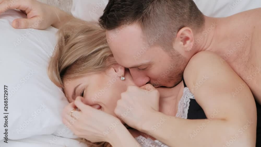 A young married couple, a man and a woman are lying on a bed with white linens. Morning and awakening. Having fun and smiling.