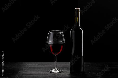 Bottle of red wine and a glass half filled with red wine, on a wooden black table, black background.