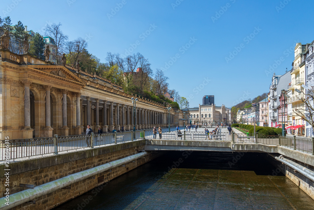 Outdoor sunny view above Tepla river, and Mill Colonnade, the largest Renaissance colonnade in town, next to promenade on riverside with full of tourists in Karlovy Vary, Czech Republic.. 