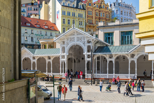 Fototapet Outdoor sunny view of tourists walk on promenade in front of Market Colonnade, Swiss style carved wooden colonnade arbour pavilion, and colorful buildings in Karlovy Vary, Czech Republic