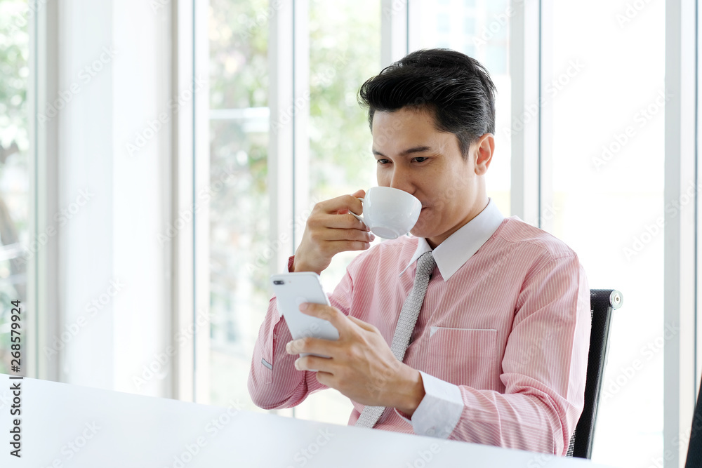 Young asian businessman using smart phone and drinking coffee at office, office life and business concept Young asian businessman using smart phone and drinking coffee at office, office life and business concept