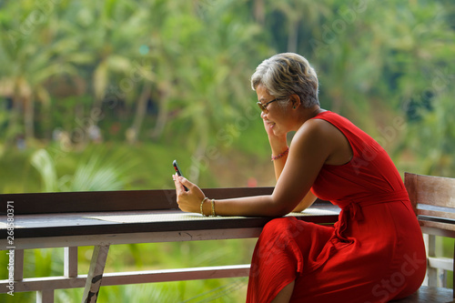 attractive and relaxed 40s or 50s Asian woman with grey hair and stylish red dress using social media on inernet mobile phone at beautiful tropical jungle resort photo