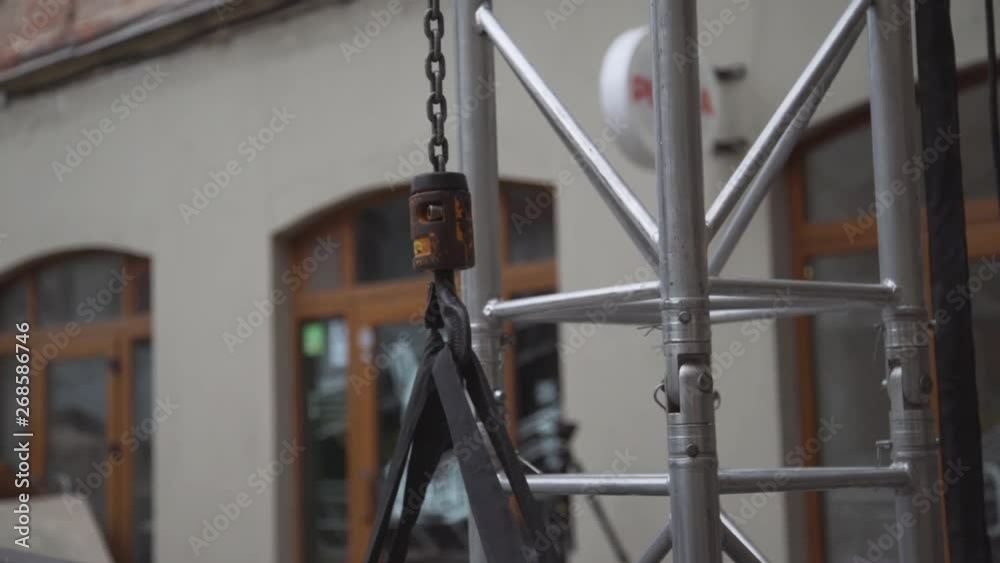 View of fixating hook connected to chain to some stage substructure located next to beige house with wooden framed windows outside at daylight on warm day. View of fixating hook connected to chain to some stage substructure located next to beige house with wooden framed windows outside at daylight on warm day.