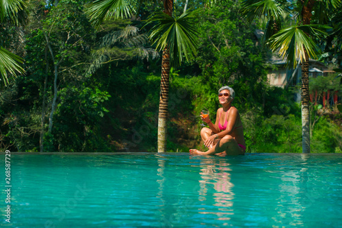 natural lifestyle portrait of attractive and happy middle aged Asian woman relaxed at tropical resort infinity simming pool with jungle background enjoying a drink relaxed