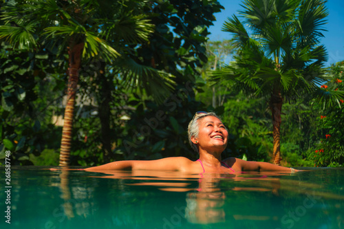  lifestyle portrait of attractive and happy middle aged Asian woman relaxed at tropical resort infinity swimming pool with jungle background enjoying relaxed