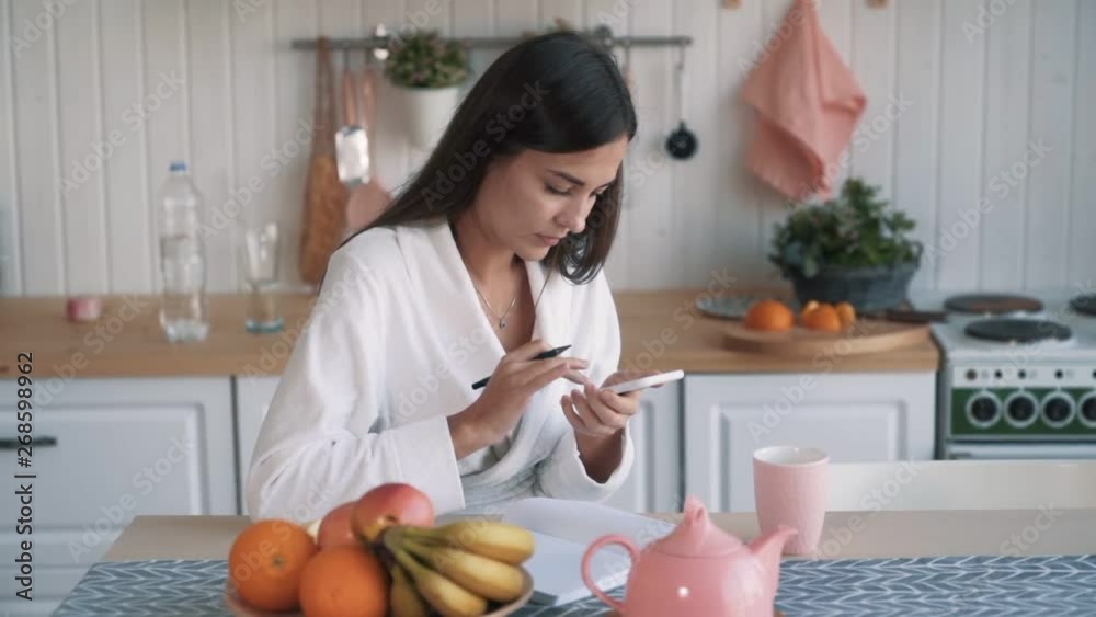 Woman uses phone, sits on kitchen and writes to do list in notebook, slow motion