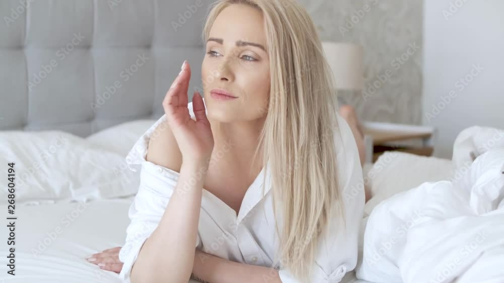 Beautiful middle aged woman lying on the bed, wearing white shirt and smiling Beautiful middle aged woman lying on the bed, wearing white shirt and smiling