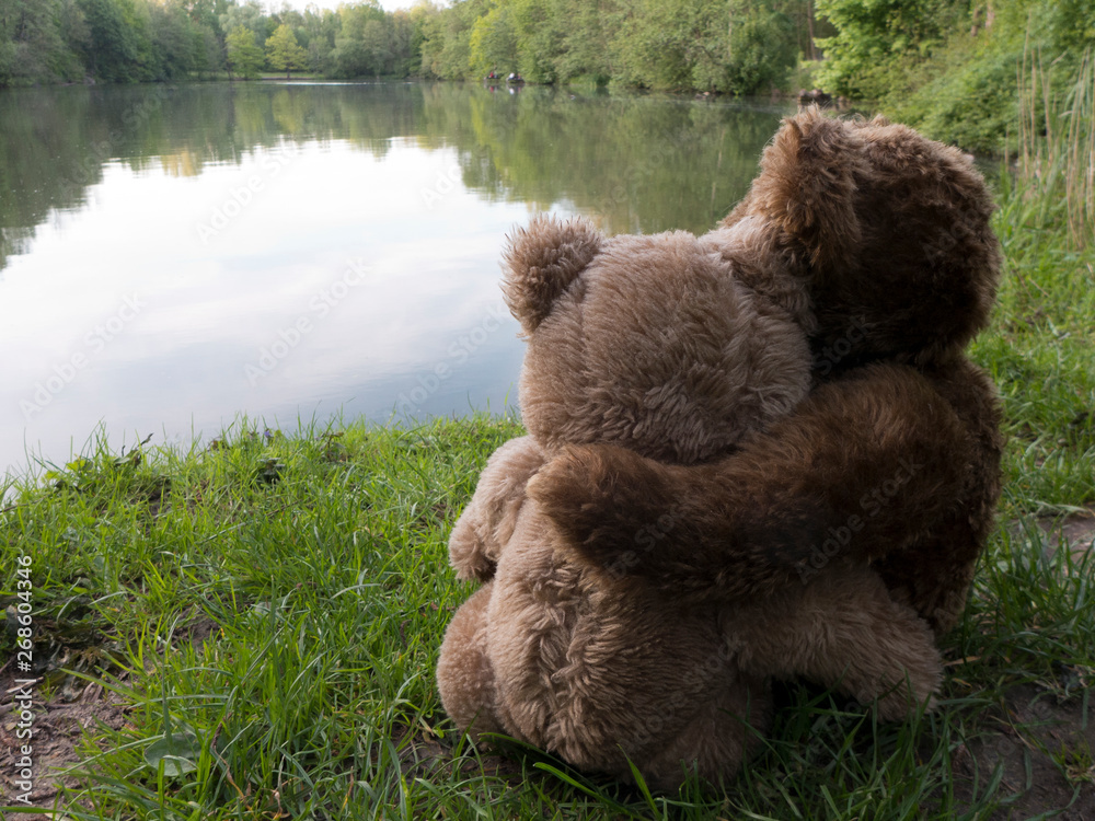 Zwei Teddybären sitzen Arm in Arm an einem See. Es ist ein romatischer Abend.