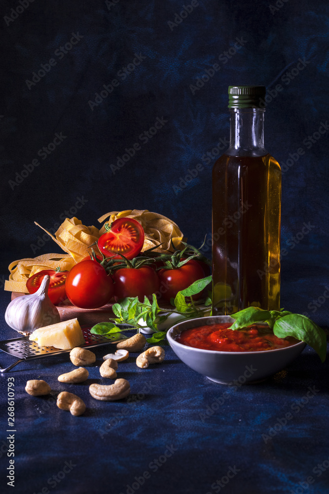 Table with ingredients to make tomato pesto. Tomatoes, garlic, fresh oregano and basil herbs, bottle of olive oil, few cashew nuts, parmesan cheese, cheese grater, wooden mortar and homemade pasta. Table with ingredients to make tomato pesto. Tomatoes, garlic, fresh oregano and basil herbs, bottle of olive oil, few cashew nuts, parmesan cheese, cheese grater, wooden mortar and homemade pasta.