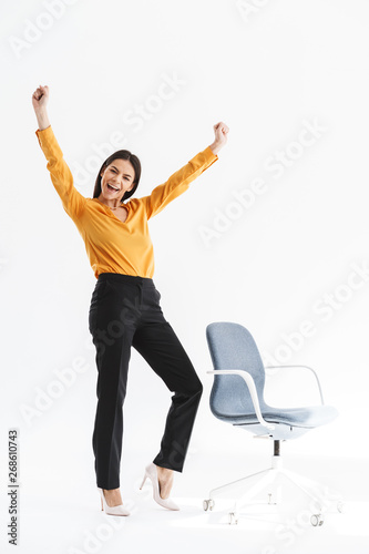 Full length portrait of ecstatic young businesswoman dressed in elegant wear rejoicing and standing by chair