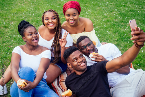 group of five friends female and male taking selfie on camera smartphone and having fun outdoors lifestyle near lake,eating burger picnic in the park