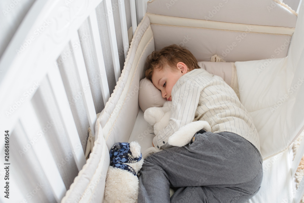 top view of a blond seven years boy sleeping in his sister white wooden cot in white shirt and grey trousers with toys in his arms top view of a blond seven years boy sleeping in his sister white wooden cot in white shirt and grey trousers with toys in his arms