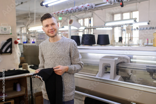 a young man analyzing a knitted piece of cloths near industrial knitted machines with black thread in cones