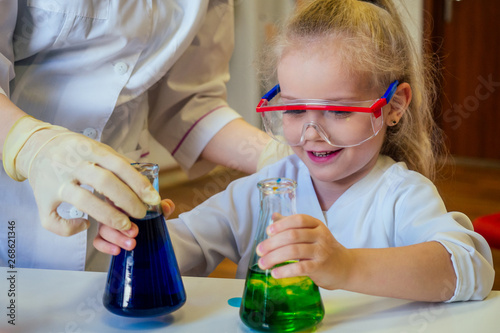 surprised and amazed clever blonde school girl chemist in eyeglasses and white medical gown making science experiments chemistry ,mixing different chemical solutions in laboratory classroom