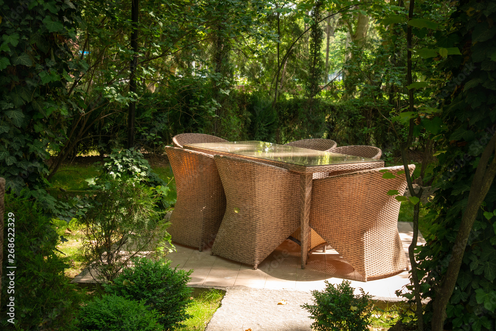 Wicker chairs and table are in the garden near trees. Wicker chairs and table are in the garden near trees.