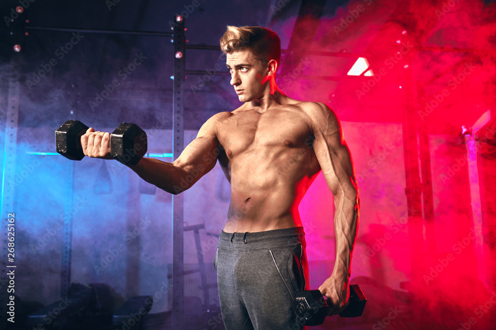 Shirtless white man in sweatpants starting exercise with dumbbell weight in dark gym with blue and red sportlight. Fitness motivation and muscle training concept.