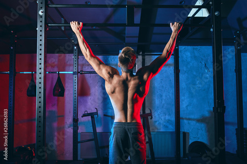 Young bodybuilder training in the gym exercising Wide-Grip Pull-Up, back view. The session covers the kipping pull-up, the butterfly pull-up and the butterfly chest-to-bar pull-up. © alfa27