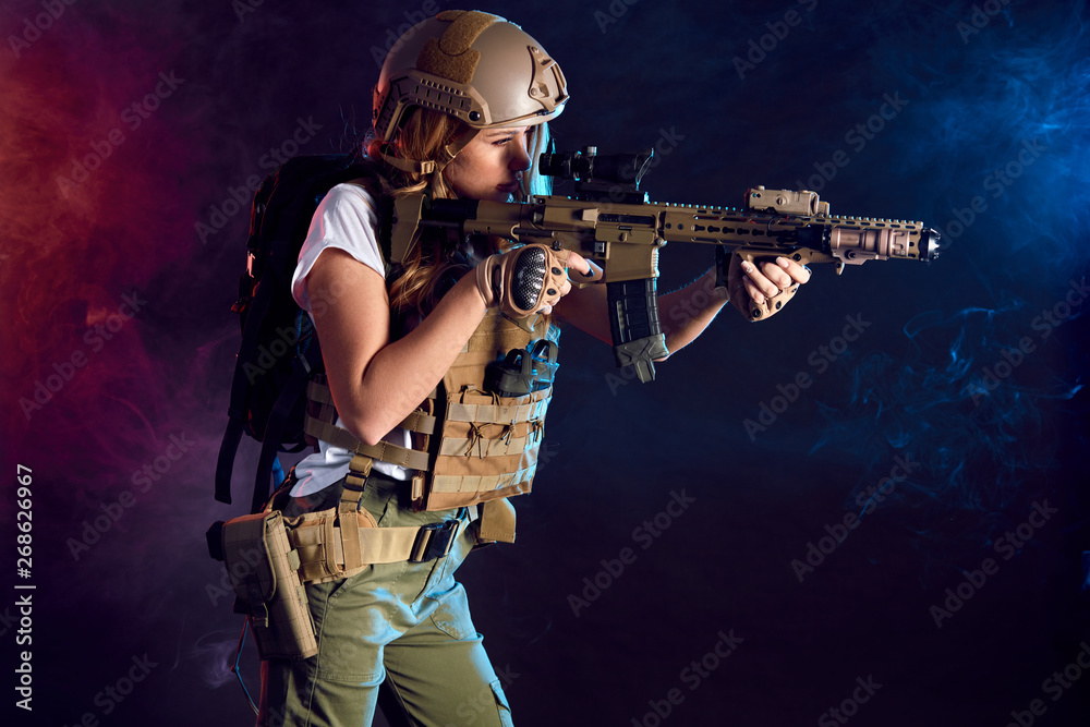 Heavily armed and well-equipped female soldier in battle helmet holding assault rifle isolated on dark smoky battlefield. Paint ball and laser tag sport games