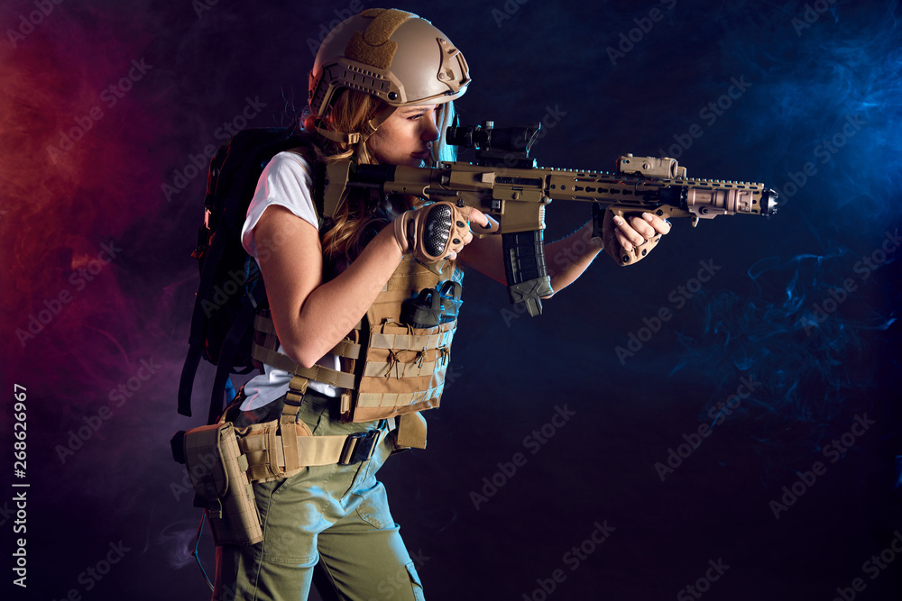 Obraz premium Heavily armed and well-equipped female soldier in battle helmet holding assault rifle isolated on dark smoky battlefield. Paint ball and laser tag sport games