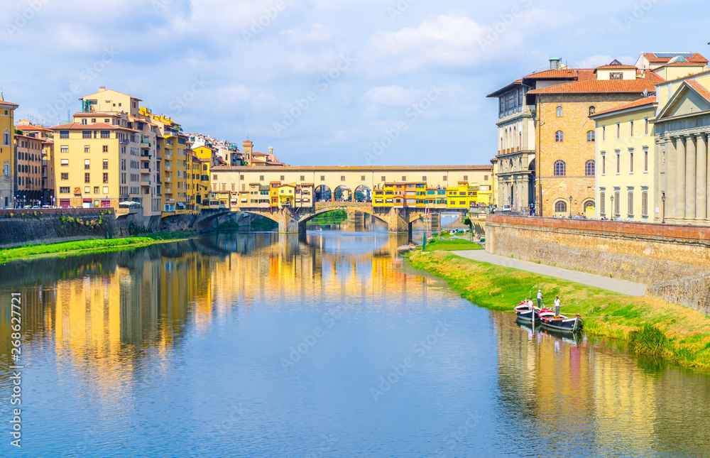 Ponte Vecchio bridge with colourful buildings houses over Arno River blue reflecting water and boats near river bank in historical centre of Florence city, blue sky white clouds, Tuscany, Italy