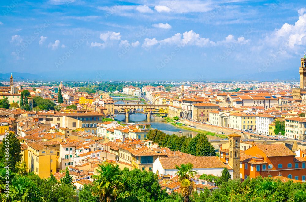 Top aerial panoramic view of Florence city with Ponte Vecchio bridge over Arno river, buildings with orange red tiled roofs, palm trees, blue sky white clouds, Tuscany, Italy