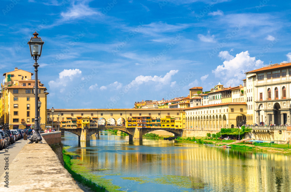 Ponte Vecchio stone bridge with colourful buildings houses over Arno River blue reflecting water and embankment promenade in historical centre of Florence city, blue sky white clouds, Tuscany, Italy