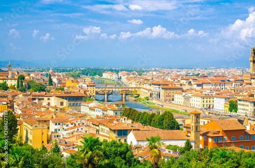 Top aerial panoramic view of Florence city with Ponte Vecchio bridge over Arno river, buildings with orange red tiled roofs, palm trees, blue sky white clouds, Tuscany, Italy