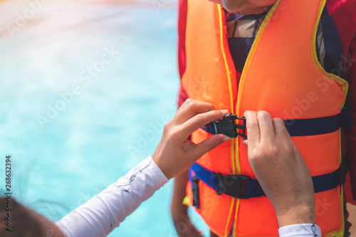 Asian mother helping her son to wear life jacket before playing in swimming pool, Selective focus.