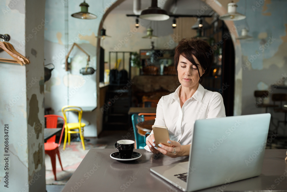 A middle-aged woman sits in a cafe, drinks coffee and works at a computer. Woman holding a phone A middle-aged woman sits in a cafe, drinks coffee and works at a computer. Woman holding a phone