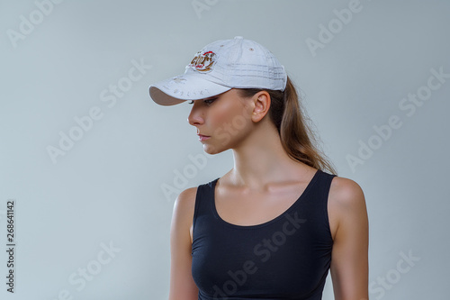 Beautiful woman in white baseball cap and black shirt posing stylishly in profile on gray background in studio. Fashion photography of accessories with sexy female model