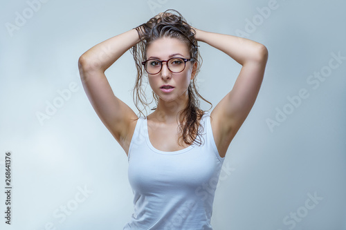 Young woman in stylish glasses and white shirt is looking at the camera and raises hair up isolated on gray background in a studio. © Alexander