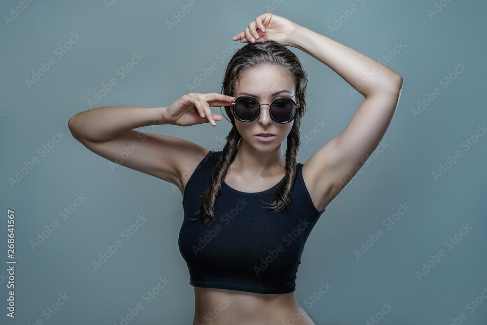 Portrait of beautiful woman in round black sunglasses and black shirt with pigtail hairstyle is posing stylishly isolated on gray background in a studio close up