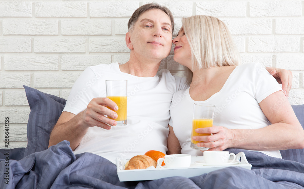 Beautiful couple having breakfast lying in bed at home. Handsome man and attractive woman are enjoying spending time together while gently hugging and kissing sitting in bed. Beautiful couple having breakfast lying in bed at home. Handsome man and attractive woman are enjoying spending time together while gently hugging and kissing sitting in bed.