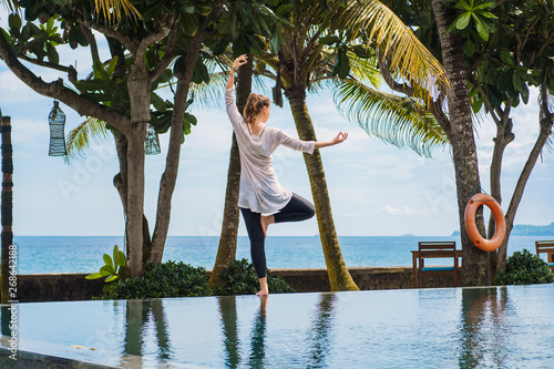 Beautiful young woman in leggings and tunic makes yoga practice, meditation, standing pose on one leg on the edge of the pool with palm trees and wonderful landscape of the ocean. Sport healthy photo © Alexander