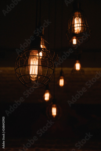 Industrial style lanterns hanged from ceiling. Metal wire lampshades in darkness. Lamps with glowing filaments inside Edison glass light bulbs. Urban style interior lighting. Wire cage lamps closeup