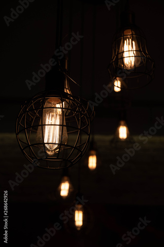 Metal wire lampshades hanged from ceiling. Lamps with glowing filament inside big glass light bulbs. Geometric lines and various carcass shapes. Urban style interior lighting. Wire chandelier closeup