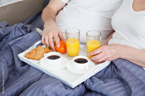 Portrait of happy playful couple relaxing in comfortable cozy bed looking at each other, cheerful man and woman having fun posing. Drink orange juice while covering their shoulders with blanket.