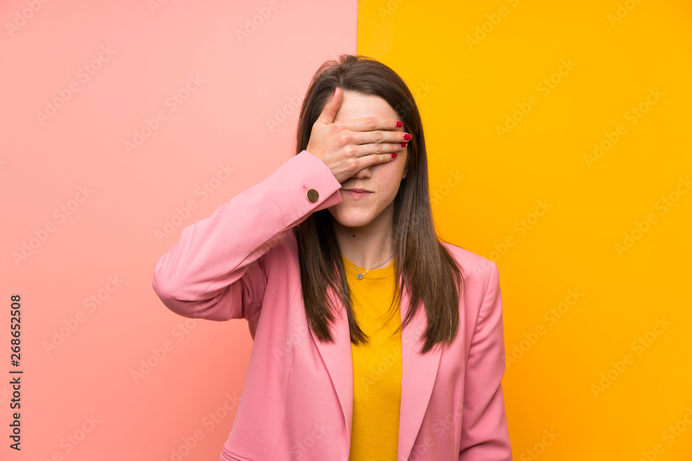 Young woman with pink suit over colorful background covering eyes by hands