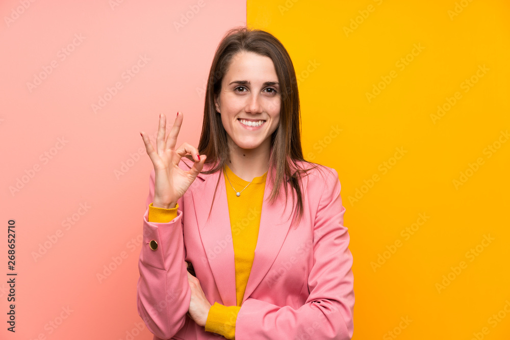 Young woman with pink suit over colorful background showing an ok sign with fingers Young woman with pink suit over colorful background showing an ok sign with fingers