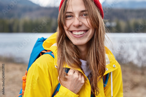 Cropped shot of smiling satisfied female tourist smiles broadly, wears yellow raincoat, has blue backpack, stands against blurred nature background, breathes fresh air, enjoys landscape outdoor © wayhome.studio 