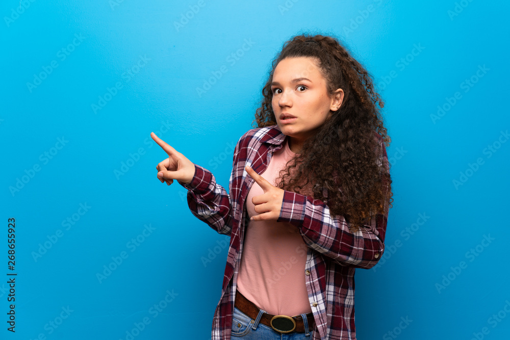 Teenager girl over blue wall frightened and pointing to the side