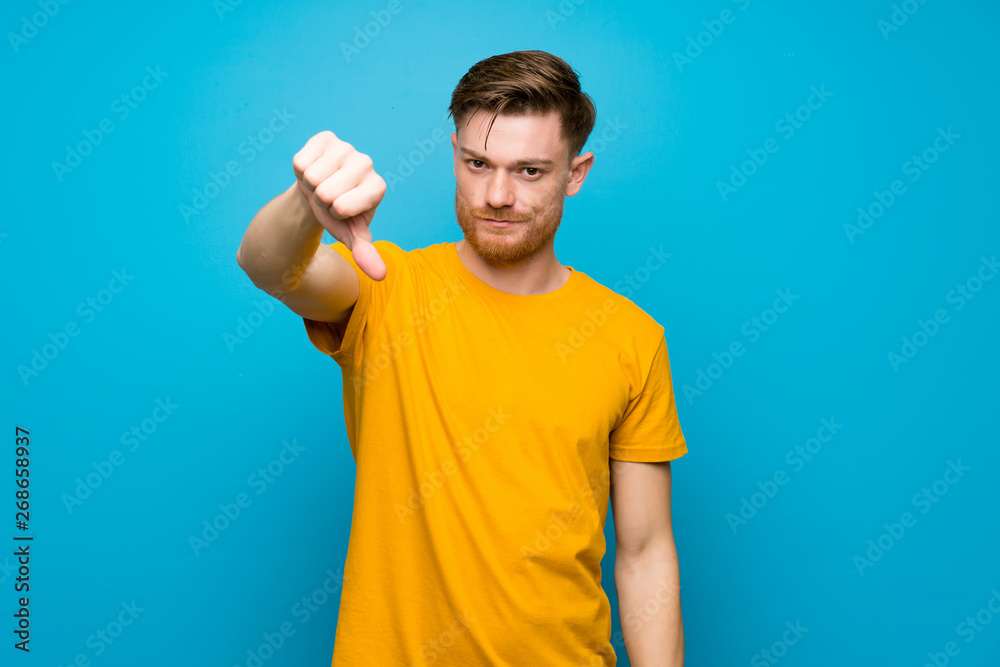 Redhead man over blue wall showing thumb down with negative expression Redhead man over blue wall showing thumb down with negative expression